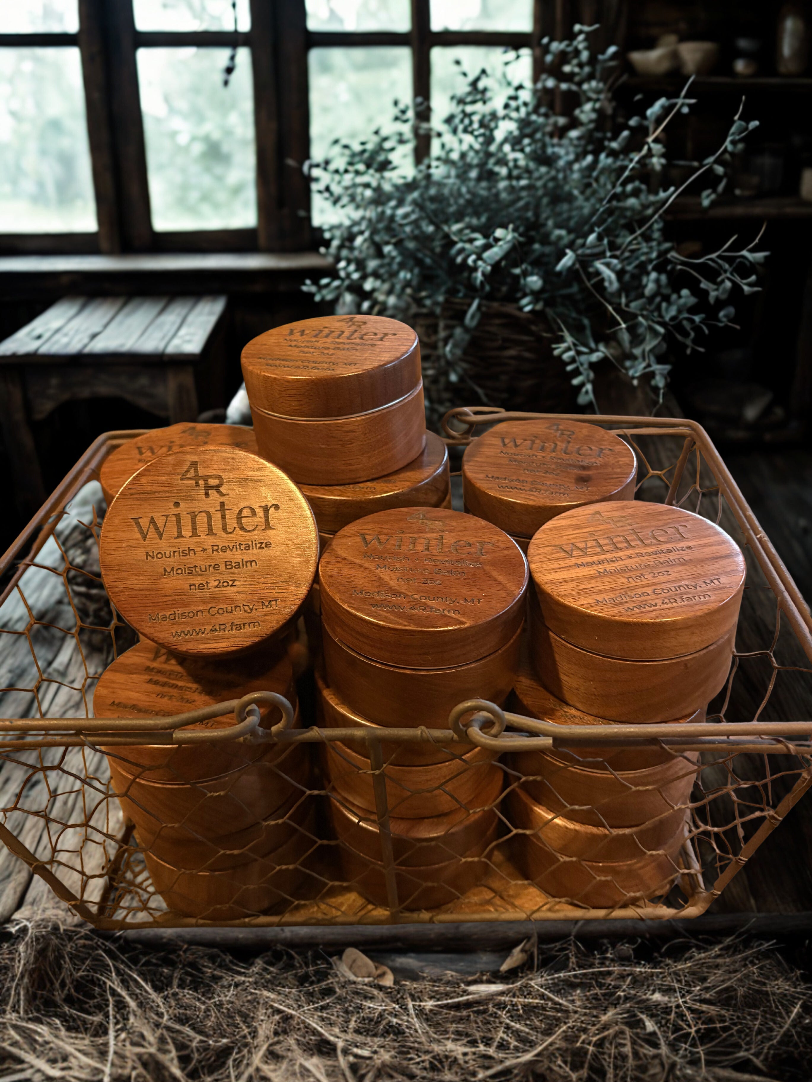 A wire basket filled with wooden jars of 4R Farm WINTER Botanical Wellness balms on a wooden table with greenery and a window
