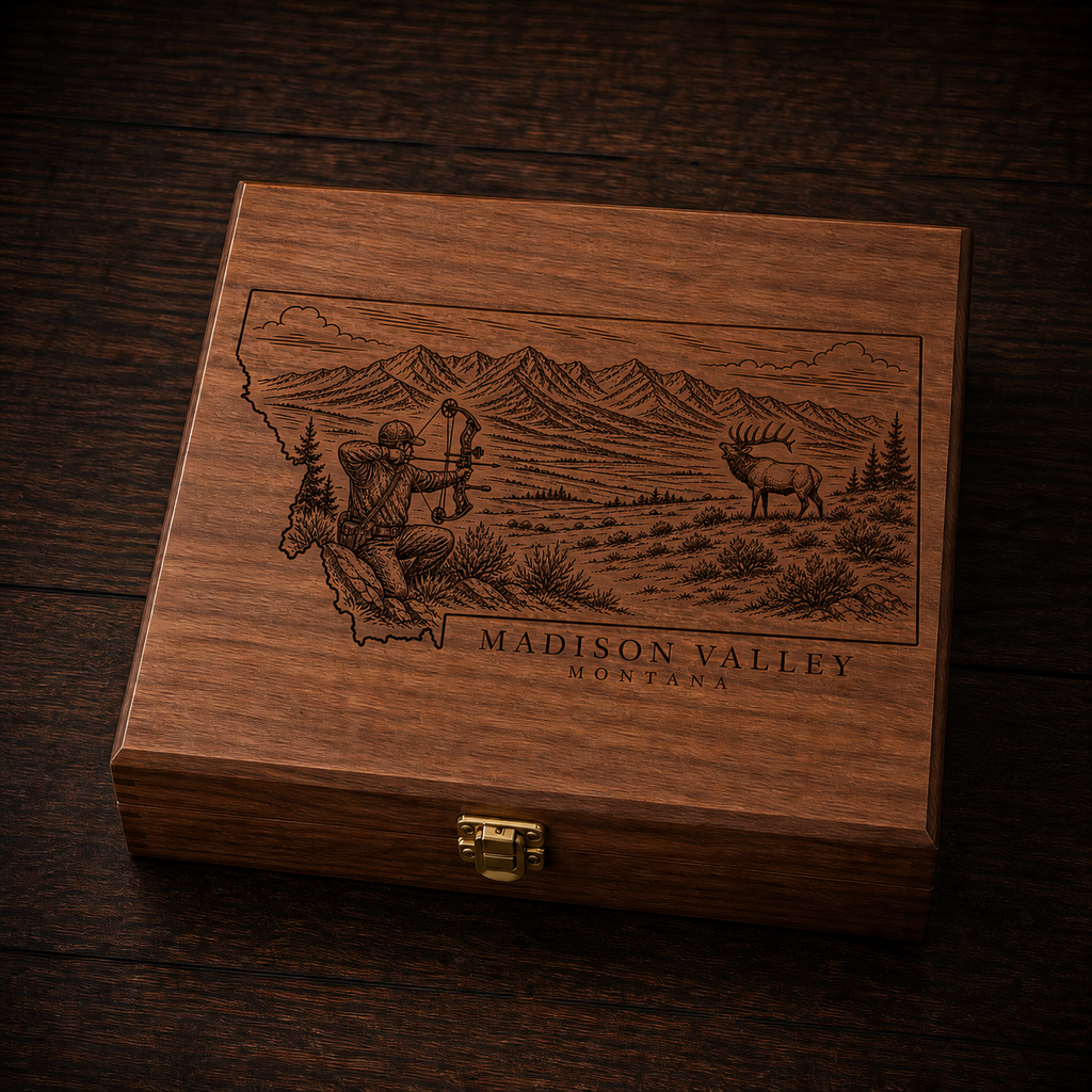 A rectangular wooden presentation box with a hinged clasp lid, featuring an engraved map of Montana with a hunter, a deer, mountains, and the text 'MADISON VALLEY HONTANA'. The box is placed on a dark wood surface.
