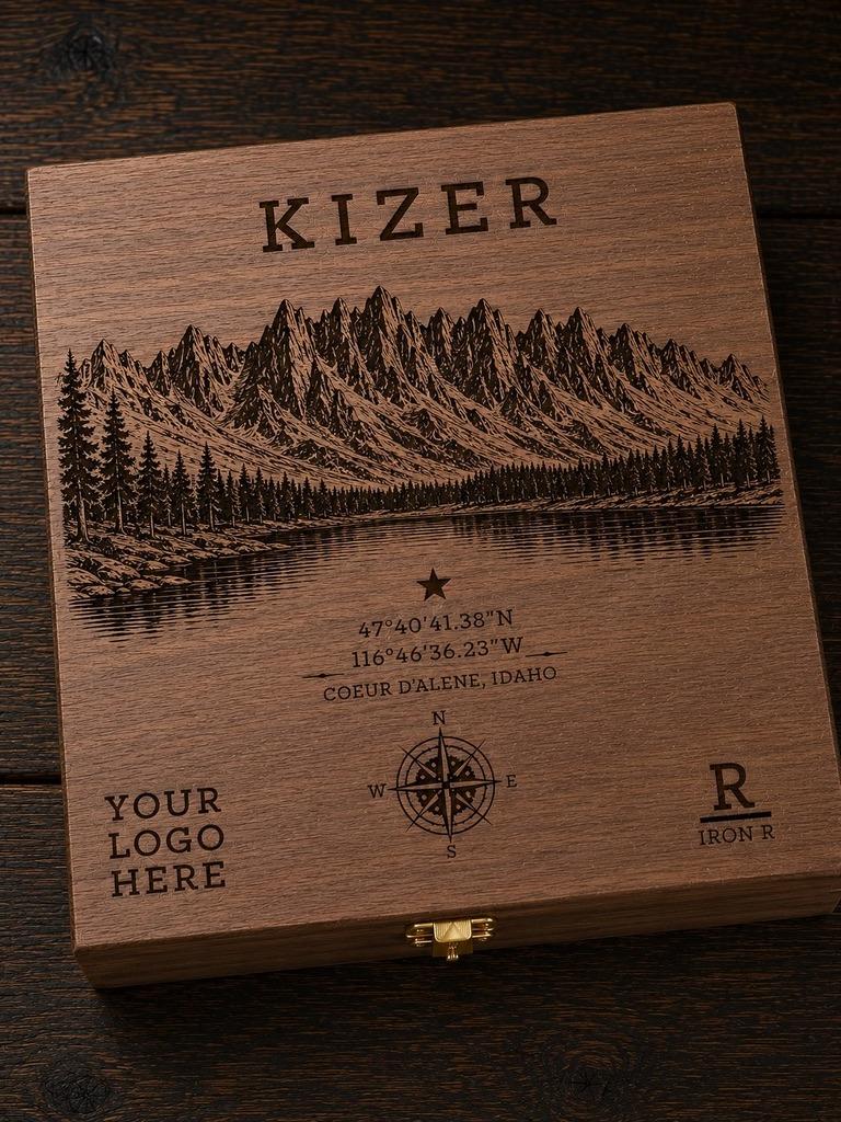 A square wooden keepsake box with a engraved mountain landscape, lake, compass rose, and customizable text including 'KIZER' and 'YOUR LOGO HERE', photographed on a dark wood surface.