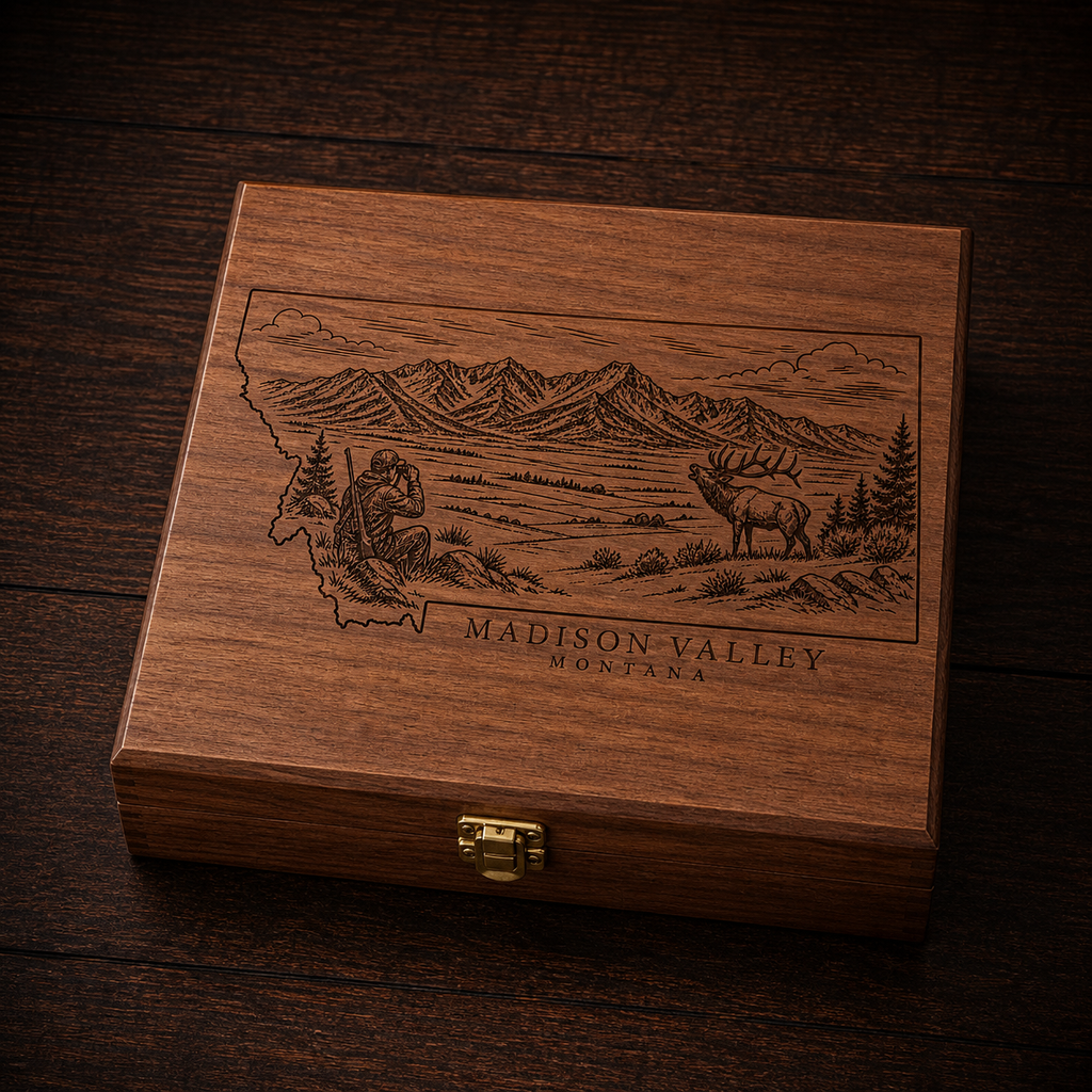 A rectangular wooden gift box with a hinged clasp lid, featuring an engraved map outline of Montana with a mountain landscape, elk, and the text 'MADISON VALLEY MONTANA.' The box is photographed on a dark wood surface.