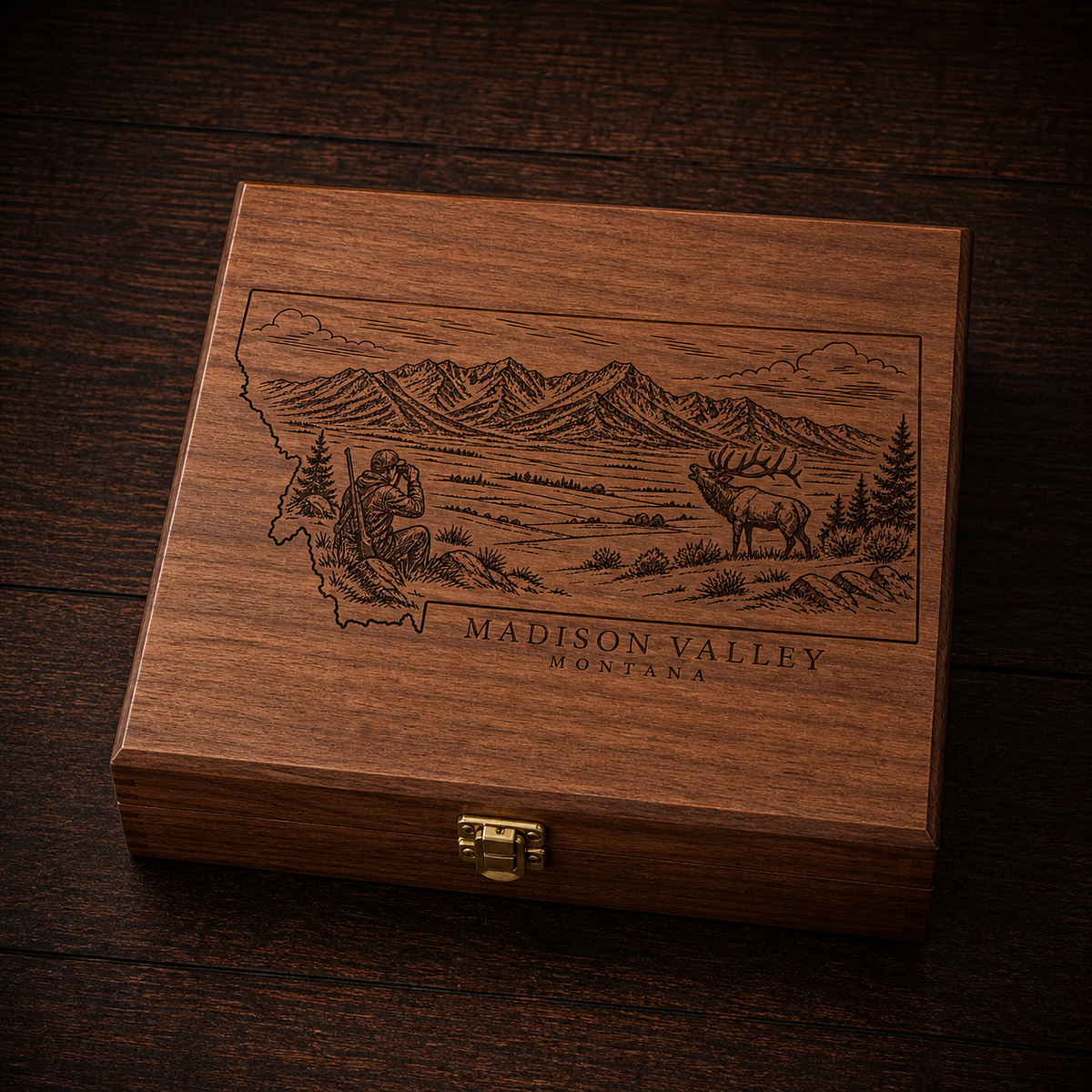 A rectangular wooden gift box with a hinged clasp lid, featuring an engraved map outline of Montana with a mountain landscape, elk, and the text 'MADISON VALLEY MONTANA.' The box is photographed on a dark wood surface.