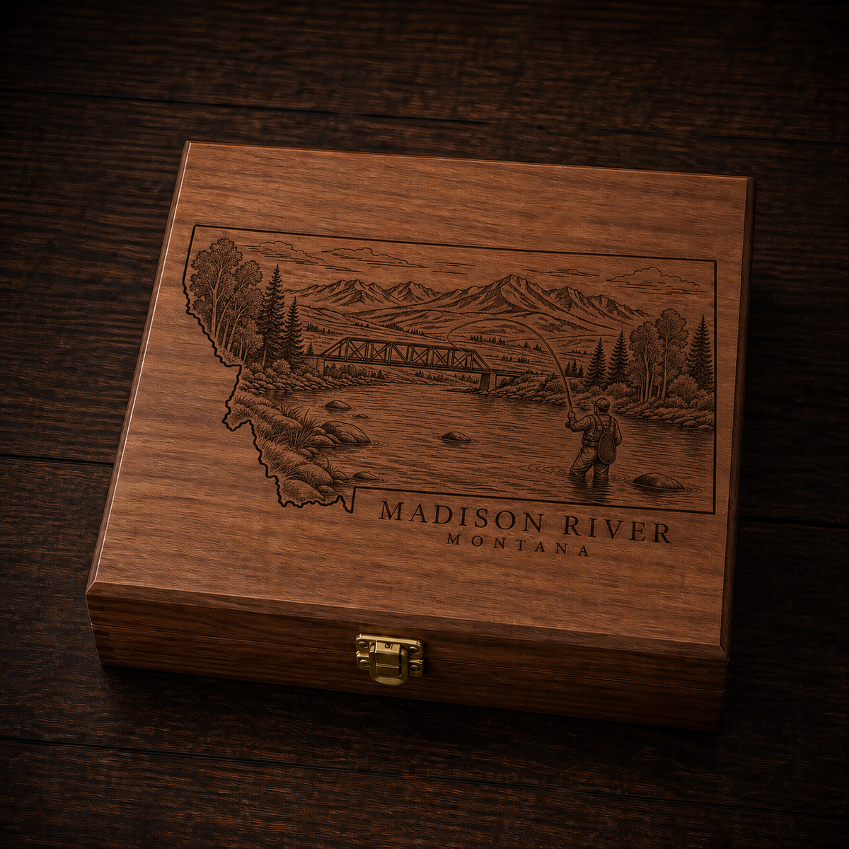A rectangular wooden keepsake box with a hinged clasp lid, featuring an engraved illustration of the Madison River in Montana with a fisherman and mountains. The box is photographed on a dark wood surface.