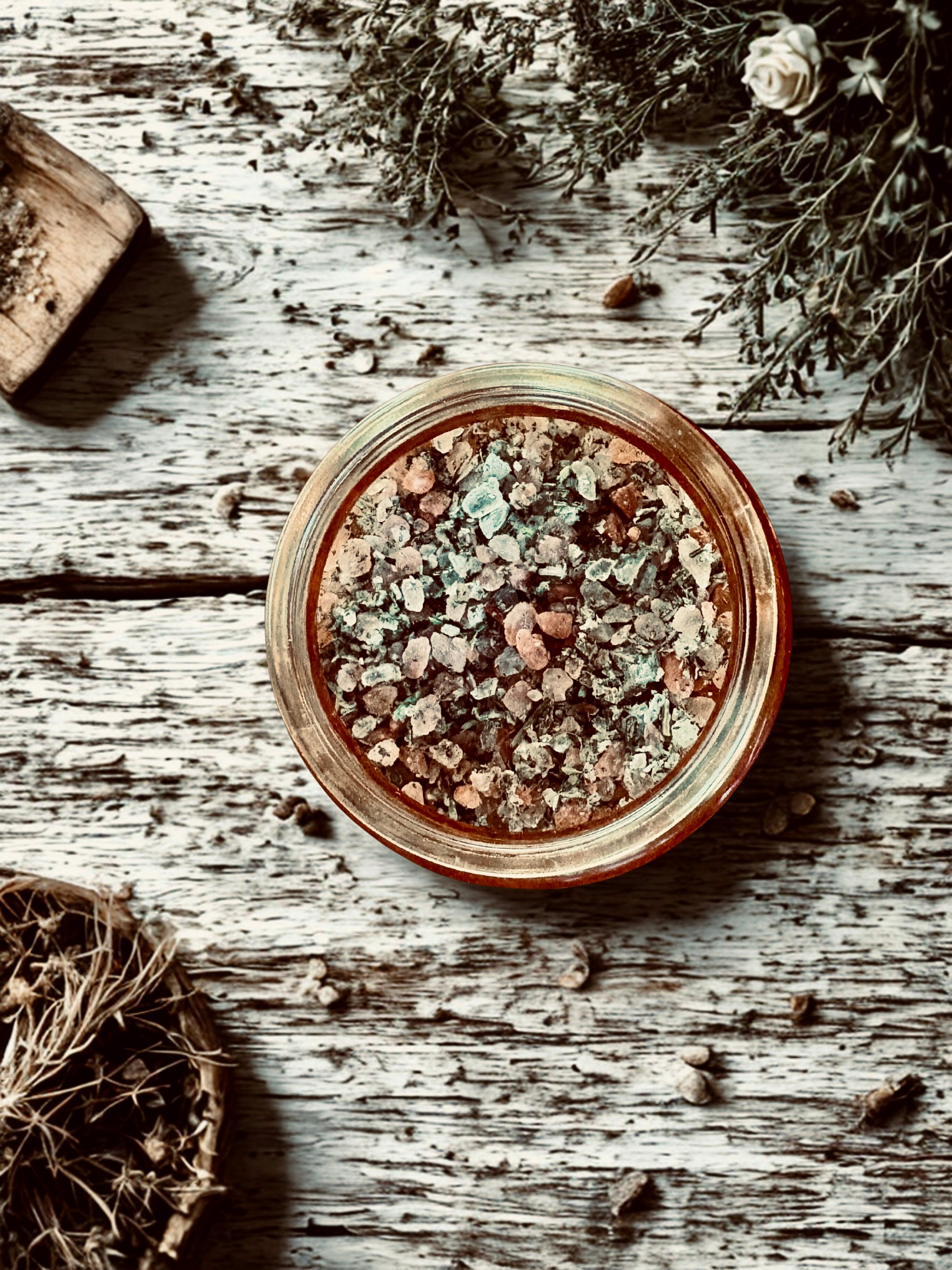 Jar of salts and herbs surrounded by dried herbs on a wooden surface with flowers and wood pieces.