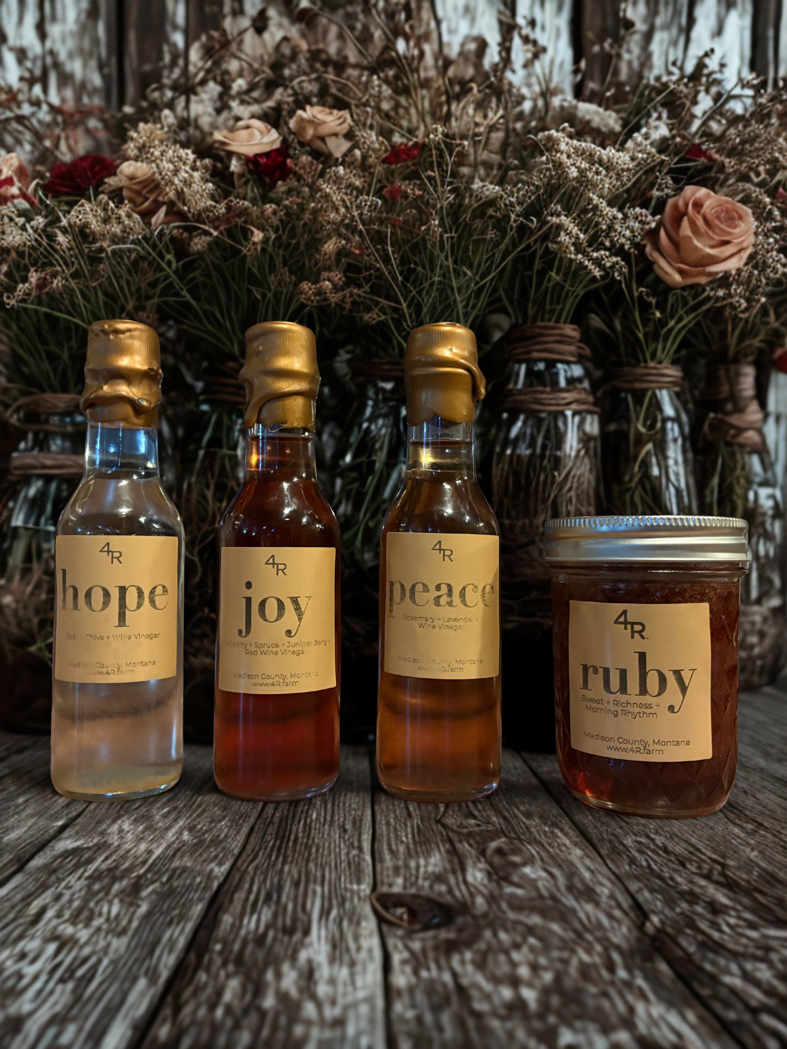Four bottles and a jar labeled 'hope', 'joy', 'peace', and 'ruby' on a wooden surface with floral arrangements in the background.